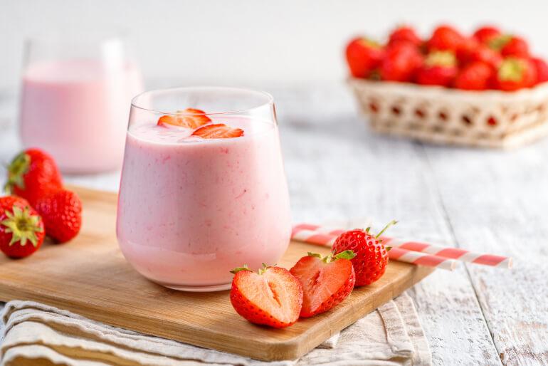 A small glass of protein yogurt smoothie sitting on a chopping board on a table surrounded by chopped strawberries