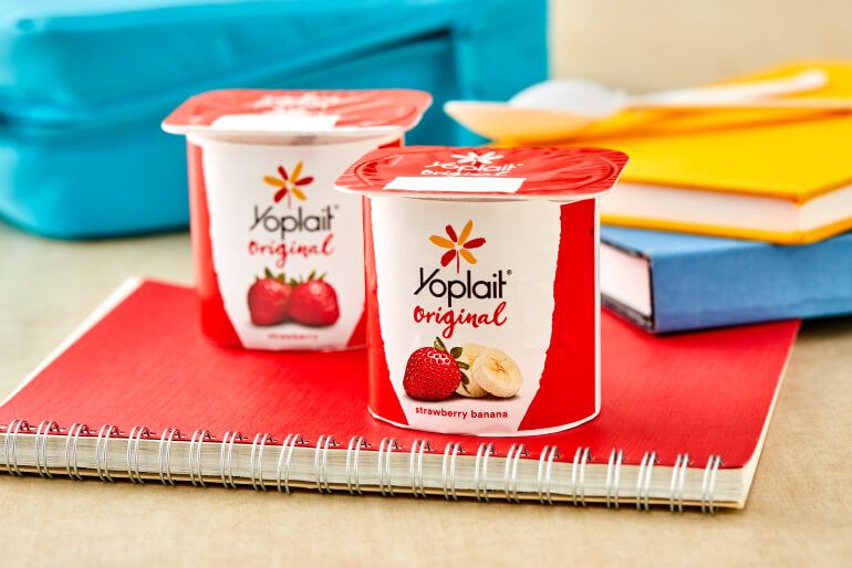 Two tubs of Yoplait Original yogurt sitting on a red notepad on a table with blue and yellow notepads behind it.